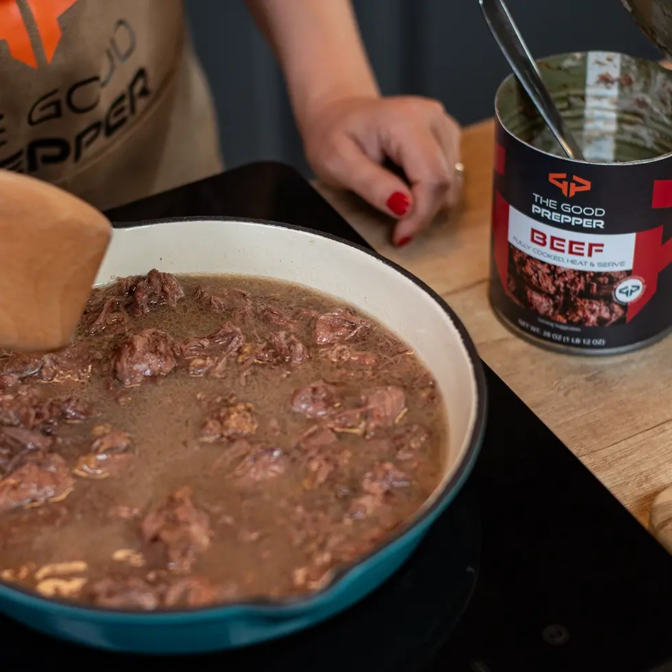 A person cooking canned beef in an iron pan on a portable stove
