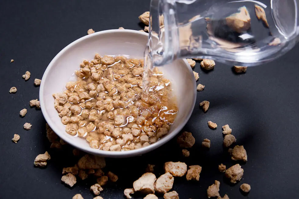 Dried raw textured soy protein (vegan meat) thin and thick, with someone's hand adding water from a jug and rehydrating on black background
